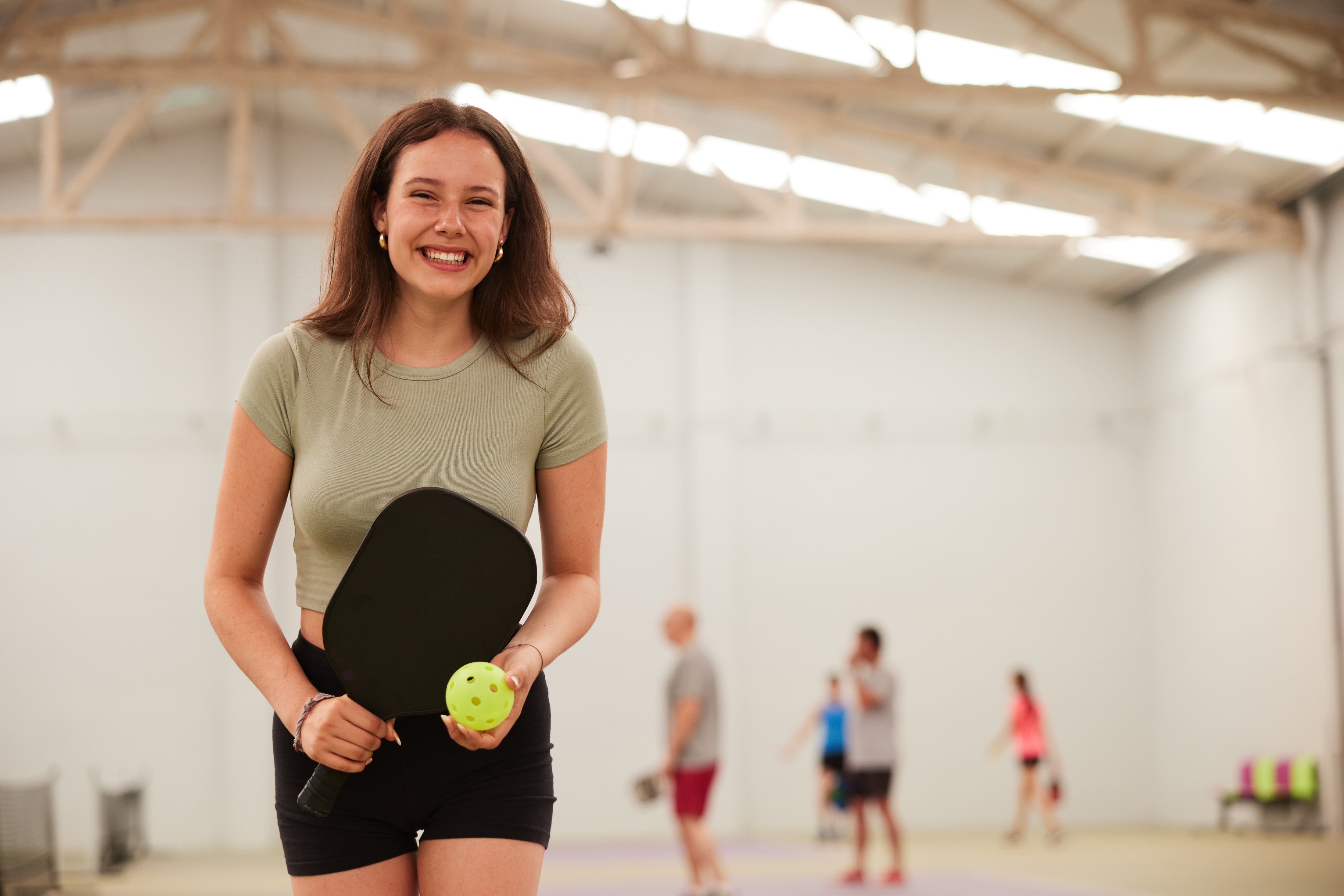 A girl in a gymnasium holding paddle and a pickleball 